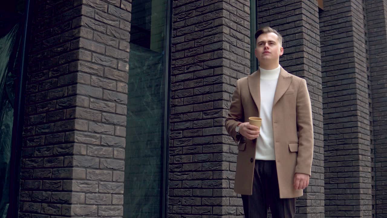 Handsome businessman in brown coat drinking coffee near the office