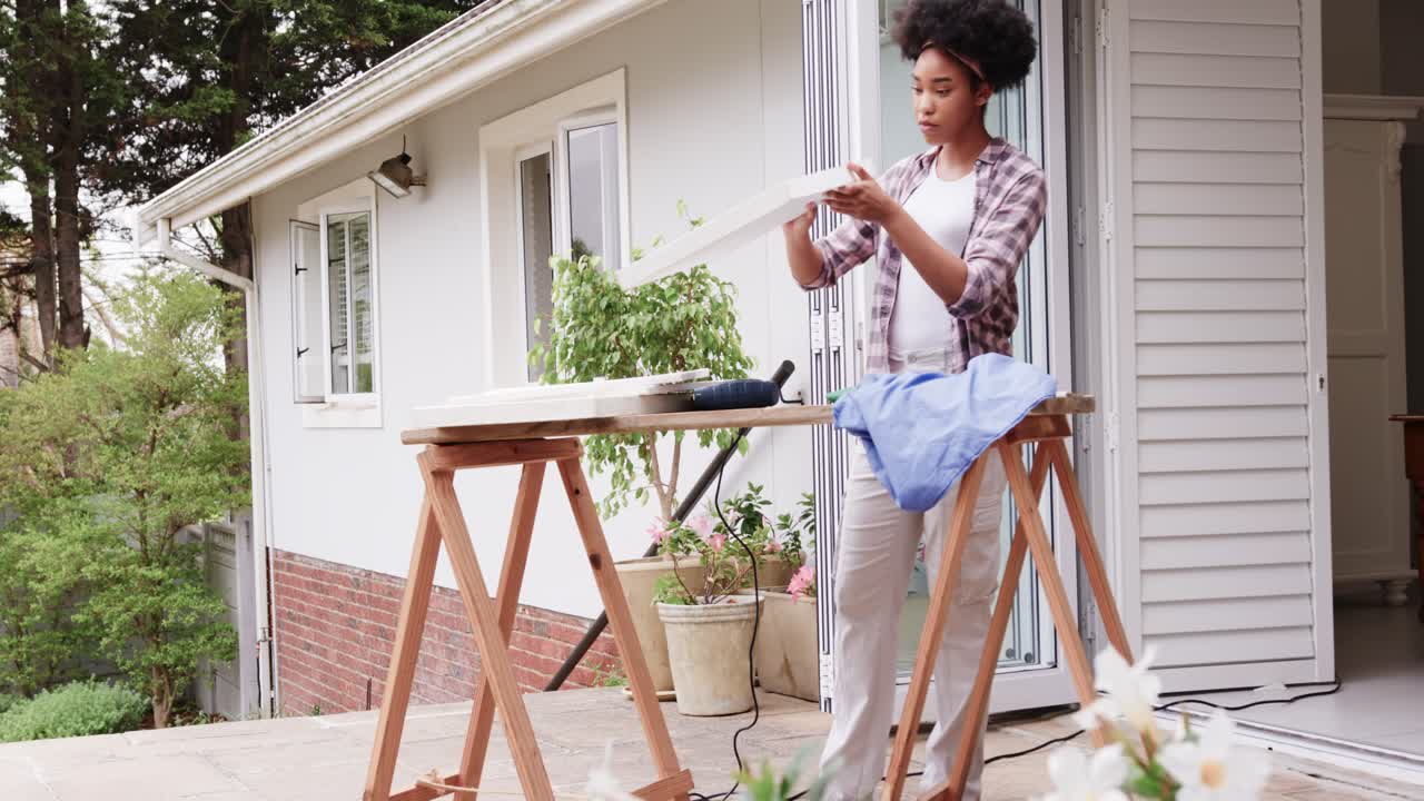 African American woman lifting panel on sawhorses on porch, checking fit for assembly, copy space