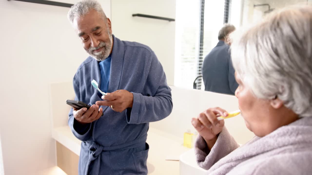 una pareja biracial feliz cepillándose los dientes, usando el teléfono inteligente en el baño, sin alterar, en cámara lenta.