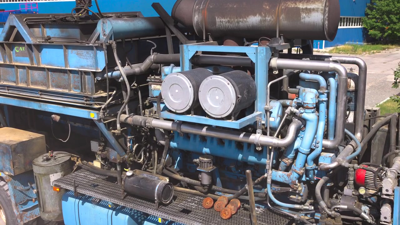 Vista laterale ravvicinata dell'assemblaggio di un motore diesel montato su camion in un piazzale industriale, che mostra il blocco motore in linea verniciato di blu, doppi filtri dell'aria cilindrici, tubi in acciaio inossidabile, tubi flessibili, valvole, dettagli della cabina e ruggine.