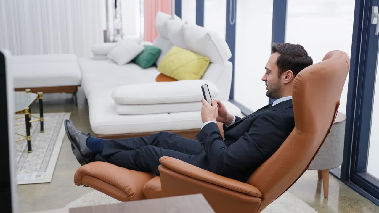 Happy businessman enjoys achievement or win by looking at phone, showing win and yes gesture while relaxing in a recliner armchair - dolly push in