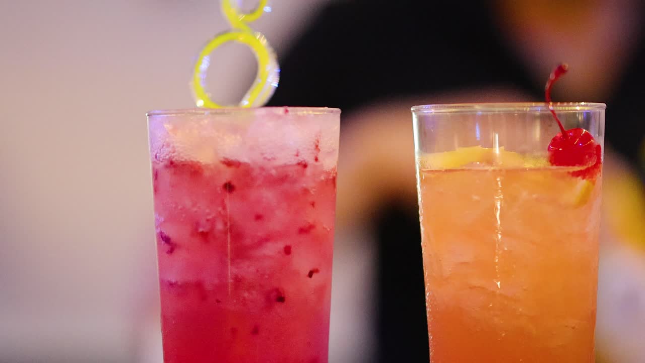 Two vibrant cocktails on bar counter, garnished with fruit, under soft, colorful ambient lighting