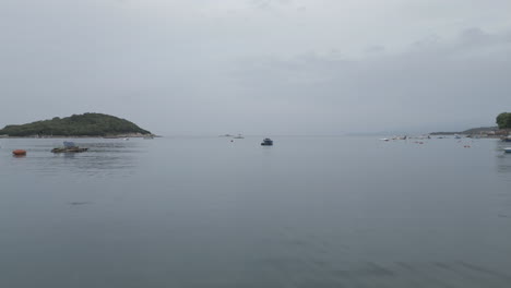 drone shot flying over the water to the small islands in front of ksamil albania in the morning on a cloudy grey day with boats around and haze in the sky log