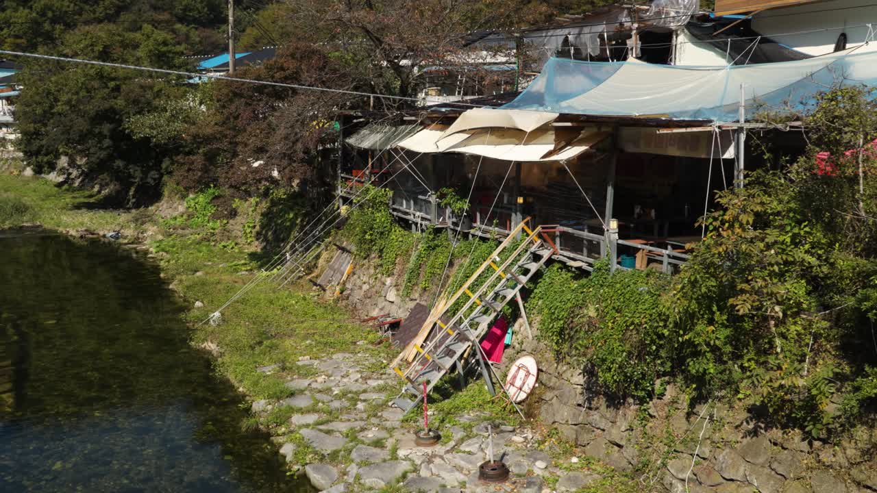A rustic traditional Korean restaurant with wooden stairs and an awning sits on a terraced stone wall beside a rocky mountain drainage canal in the natural, lush landscape of Naejangsan