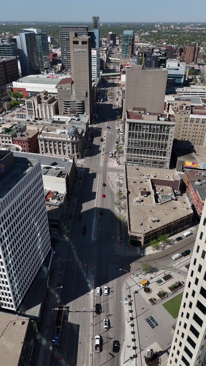 Portage Avenue nel centro di Winnipeg, Manitoba. Drone statico dall'alto guarda verso ovest lungo la strada mostrando il traffico in movimento, gli edifici cittadini e la griglia urbana in una soleggiata giornata estiva