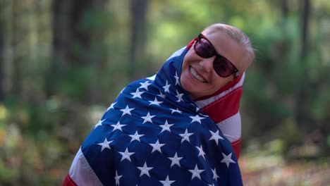 primer plano de una mujer rubia mirando a la cámara y levantando una bandera americana detrás de ella con expresión sonriente y envolviendo la bandera alrededor de su cabeza y cuerpo