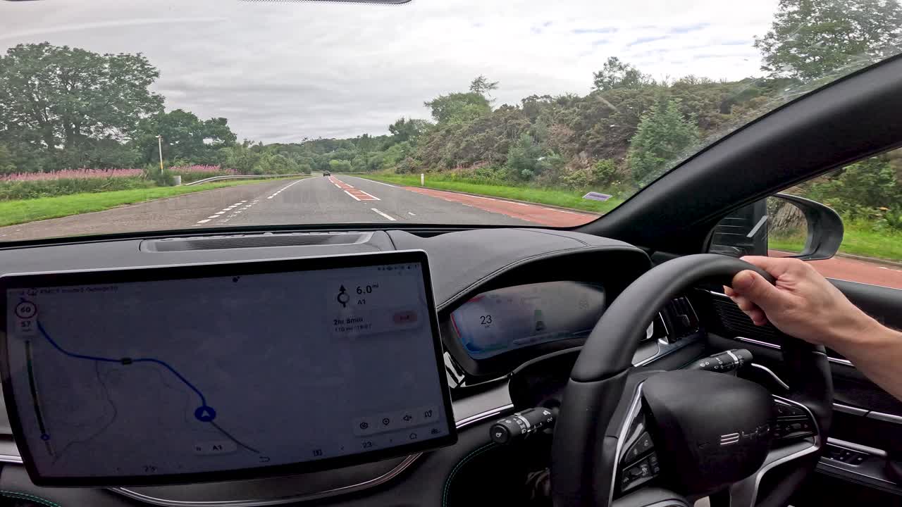 Person operates a modern car using a large touchscreen navigation system while driving along a scenic countryside road in Durham, England, under overcast daylight