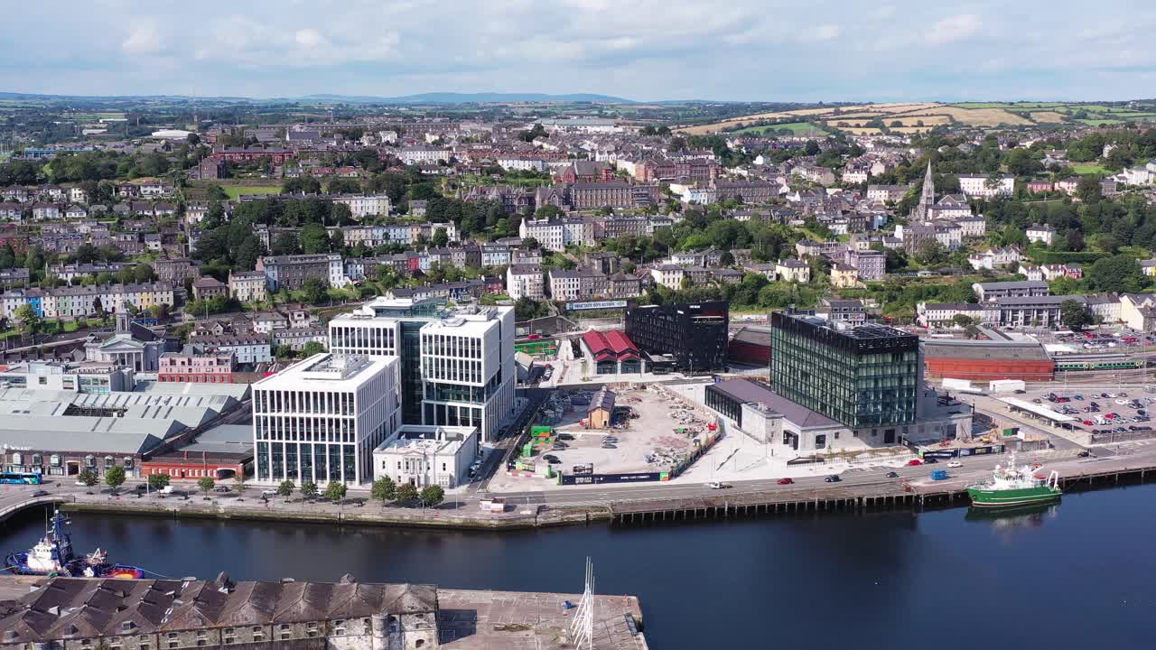 One of Ireland’s coastal cities during bright daytime. A flyover shot that pans left around part of the city of Cork, showcasing the streets and houses below along with the train station and boats.