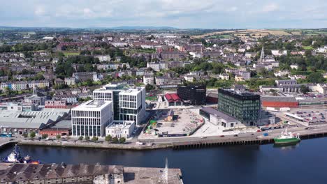 one of ireland’s coastal cities during bright daytime. a flyover shot that pans left around part of the city of cork, showcasing the streets and houses below along with the train station and boats.
