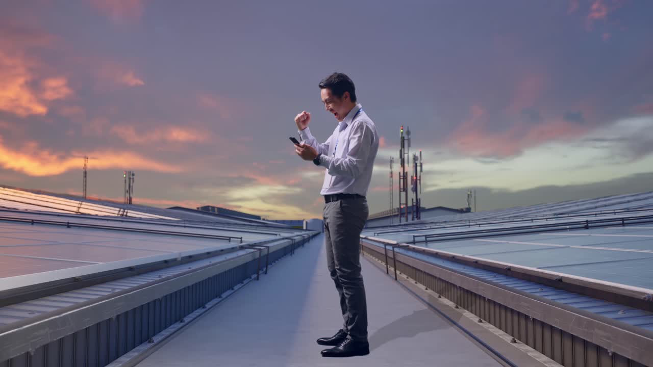 Vista laterale a figura intera di un professionista maschile asiatico in piedi con il suo telefono, mentre ispeziona pannelli solari industriali sul tetto. Alza il pugno e urla "Goal!".