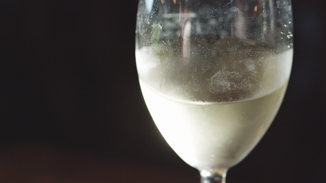 A glass of chilled white wine is gently swirled, creating visible motion and condensation. Soft, moody lighting highlights the liquid’s clarity and coolness