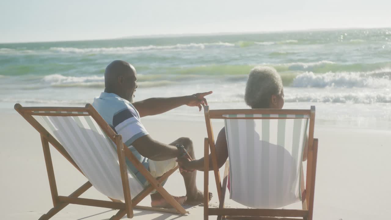 Happy senior african american couple sitting on deck chairs, holding hands at beach, in slow motion