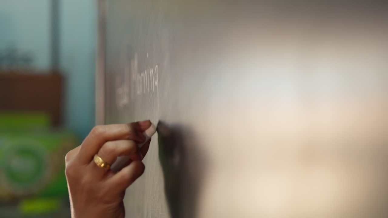 A teacher's hand writes "good morning" in English with white chalk on a blackboard in a rural Indian classroom. The hand moves steadily across the board, wearing a ring, with blurred classroom.