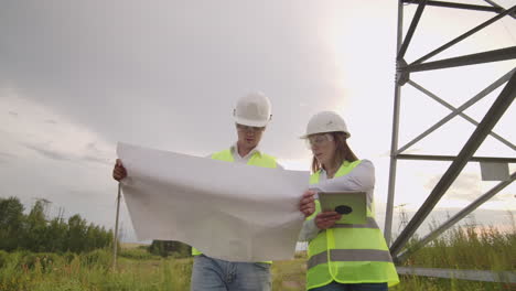 Two-electricians-work-together-standing-in-the-field-near-electricity-transmission-line-in-helmets.-Two-electricians-work-together-standing-in-the-field-near-with-power-transmission-towers.-Eco-friendly-fuel