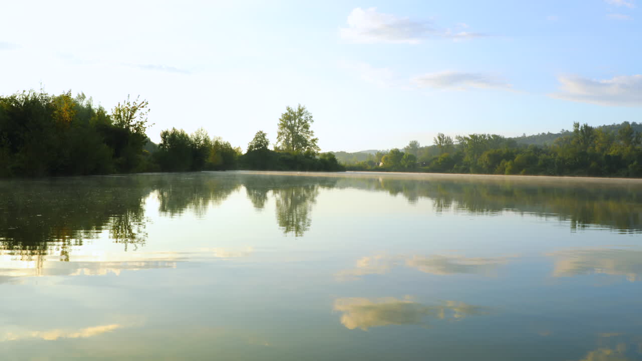 안개가 물 위로 떠오르고 태양이 탁 트인 전망 4k로 물에 반사되는 구름 뭉치와 함께 호수에 기어 다니는 나무 뒤에 빛날 때 일출의 호수 표면의 전망
