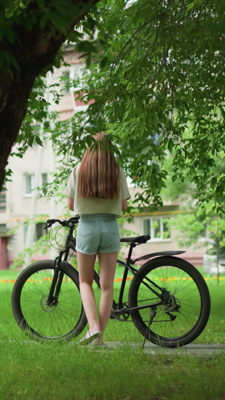 캐주얼 여름 의상을 입은 젊은 여성이 포장 된 길에 주차 된 검은색 자전거에 접근하여 좌석에 물건을 놓고, 배경에는 울창한 초록색, 높은 나무 및 주거 건물이 있습니다.