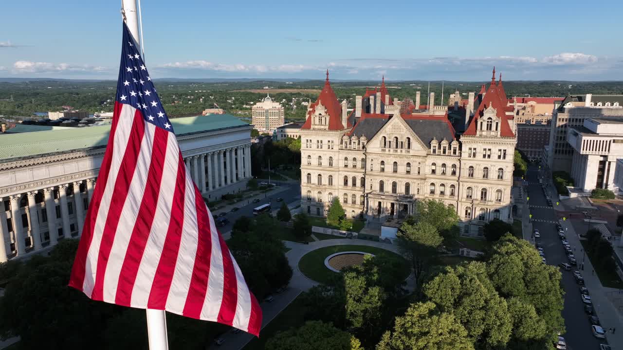 Vajende amerikansk flag på flagstang i West Capitol Park i Albany. Luftfoto tæt på. Chancellor Hall og New York State Capitol Building i det fjerne. Gylden time om aftenen. Sommersæson.