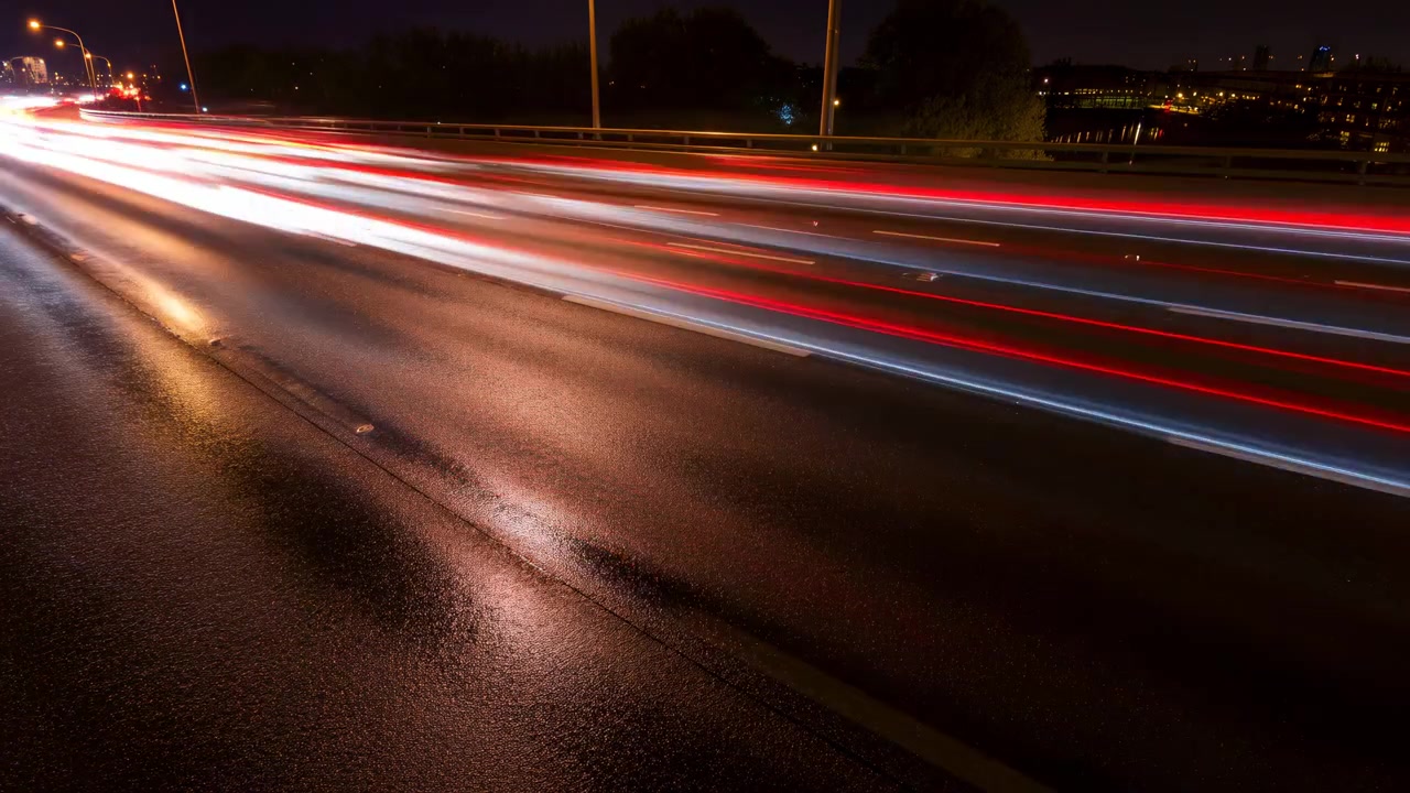 Scie luminose notturne su autostrada bagnata, fasci rossi e bianchi che attraversano l'asfalto riflettente e un cavalcavia scuro.