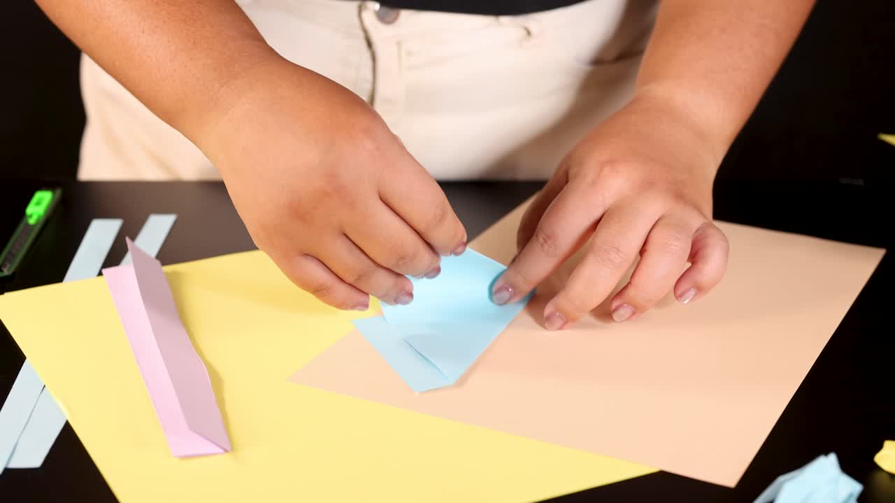 Person folds blue origami paper on desk under bright lighting, close-up, steady overhead camera