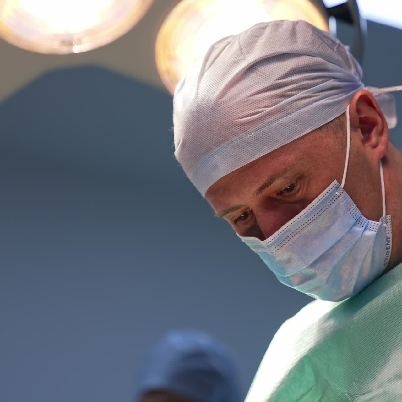 Male doctor in mask looks focused on the surgical procedure. Big lamps are on over the surgeon's head. Low angle view