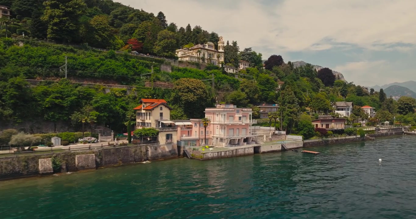 Il drone scivola lungo eleganti ville storiche sul Lago Maggiore mentre le auto percorrono la strada costiera. Acqua di un blu intenso e uno spettacolare paesaggio alpino creano un paesaggio italiano senza tempo
