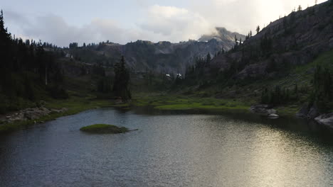 Aerial-dolly-above-alpine-lake-to-grassy-meadow-and-evergreen-trees-in-PNW-forest-with-mountain-peak-above