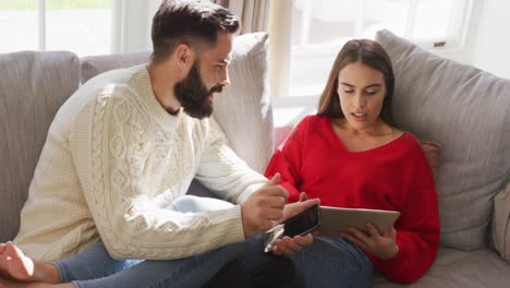 video de una feliz pareja caucásica sentada en el sofá y usando una tableta y un teléfono inteligente