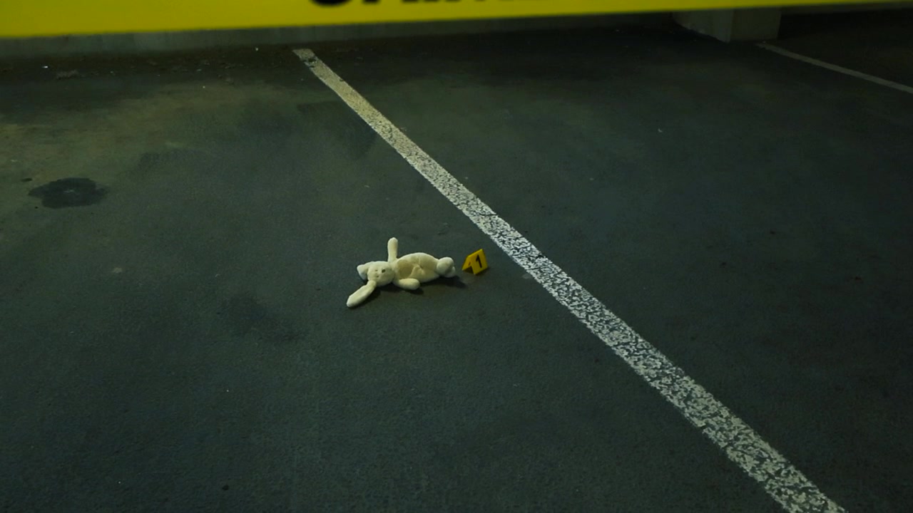 Yellow police tape reading 'Crime Scene Do Not Cross' moving to reveal a toy bunny lying on the ground next to a numbered evidence marker inside a dark underground parking garage