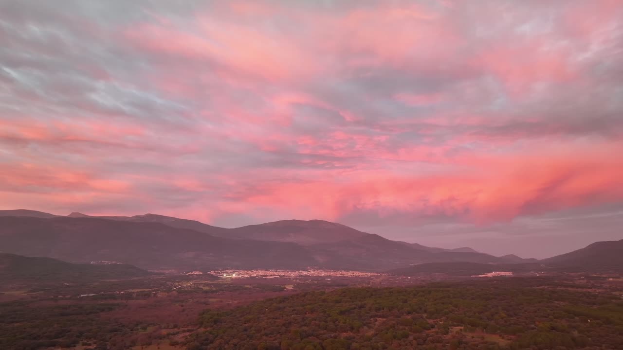 Volo laterale di un drone su una valle circondata da montagne e con diverse città al tramonto, con nuvole rosa-arancio e viola ad Avila, Spagna.