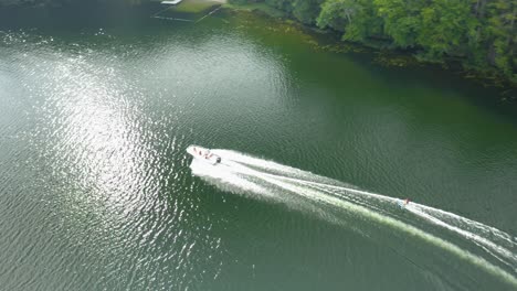 Aerial-drone-bird's-eye-view-over-water-skier-skiing-by-a-speedboat-on-sunset-lake,-New-Hampshire,-USA-during-evening-time