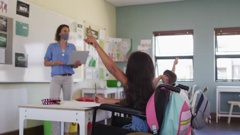 chica discapacitada con máscara facial levantando la mano mientras está sentada en silla de ruedas en clase