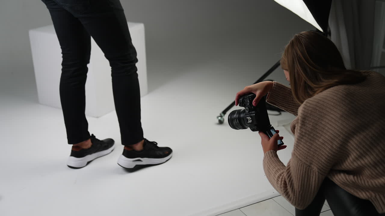 Lady taking shot of male feet in new modern black sneakers with a white sole. Backstage work of a photographer in studio.