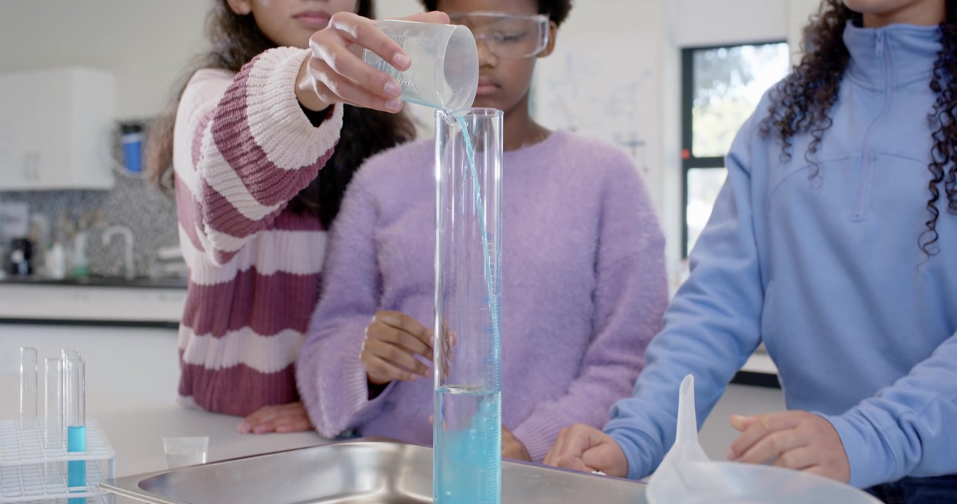 Tre giovani studentesse iniziano a misurare, versando un liquido blu in un cilindro graduato in laboratorio.