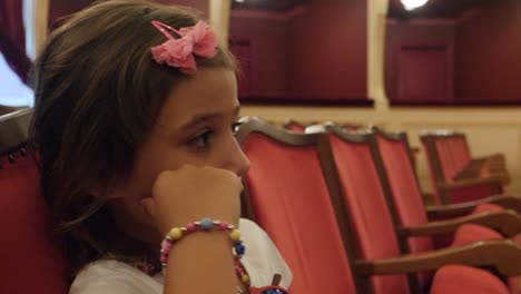 a bored little girl sitting in a theatre seat watching the screen or stage