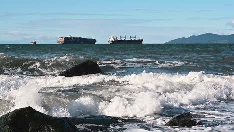 wild rooling waves from the ocean splashing on the gray rocks with on the background a giant container vessel and bulk carrier sailing on a partly cloudy day