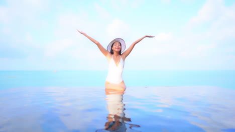 Mujer-Exótica-Feliz-En-La-Piscina-Infinita-Fusionándose-Con-El-Mar-Y-El-Cielo,-Levantando-Los-Brazos-Llenos-De-Felicidad