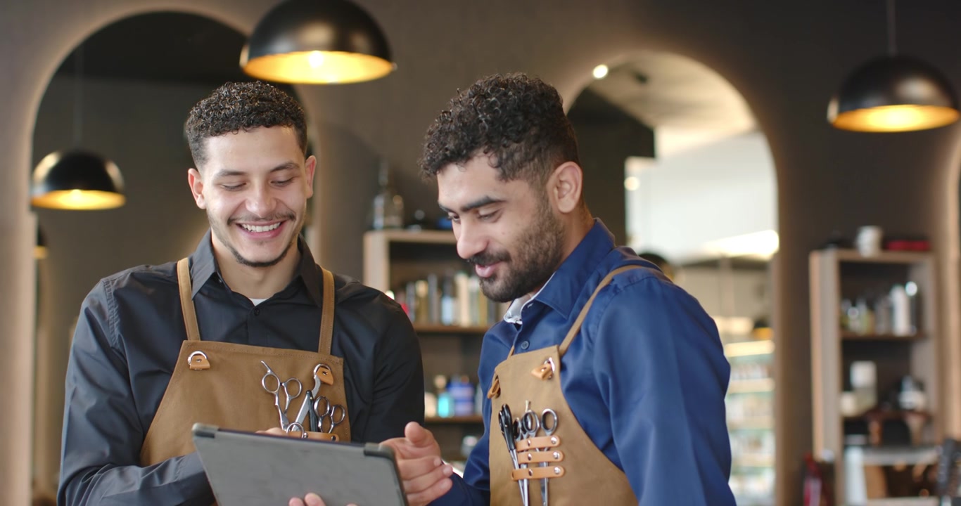 Barbeiros afro-americanos usando tablet, apontando e planejando cortes na barbearia, vestindo aventais de couro