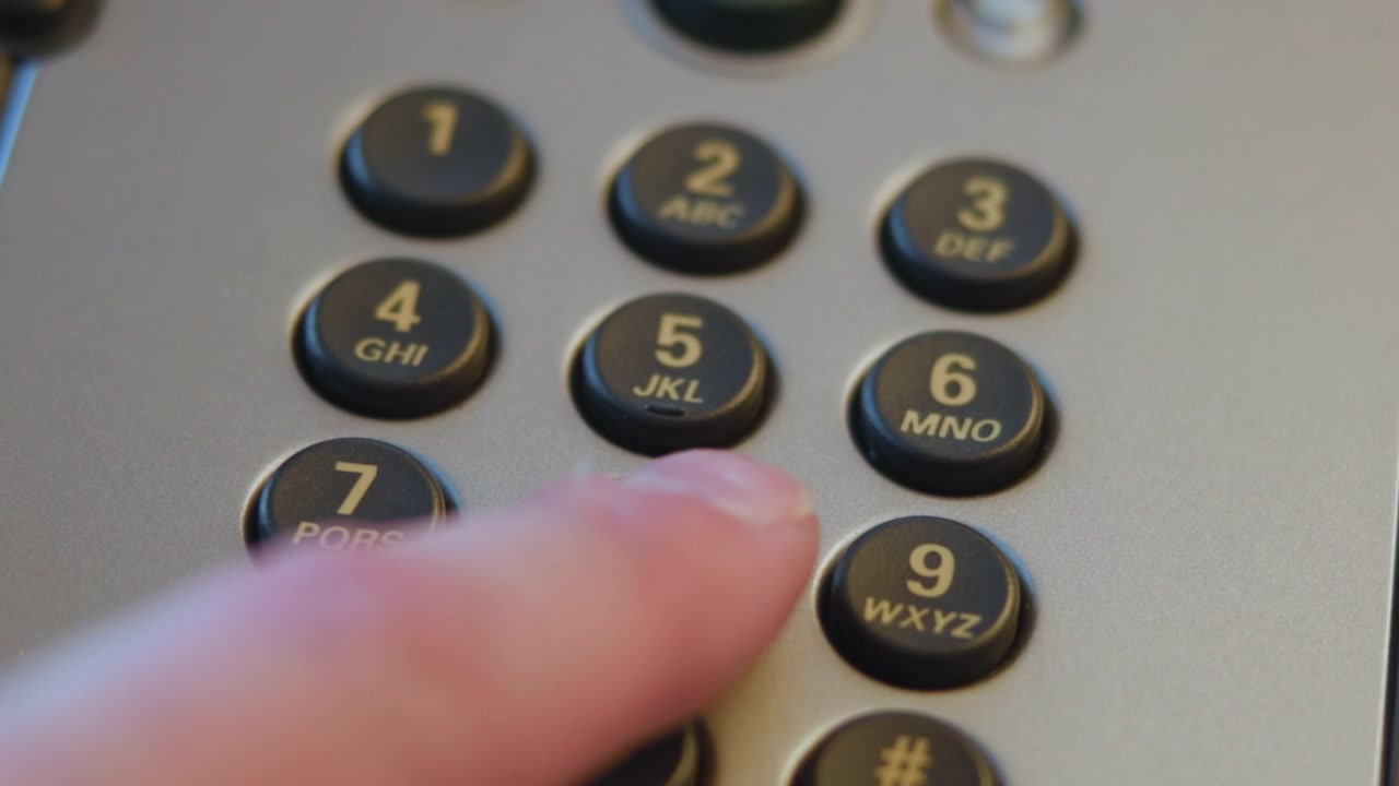 Close-up fingertip pressing numeric keypad on office desk telephone under warm diffuse light, showing tactile contact, recessed round keys, matte silver housing, shallow focus. High quality 4k footage