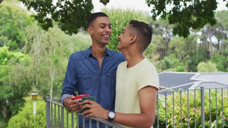 Mixed-race-gay-male-couple-standing-in-garden-drinking-cups-of-coffee-talking-and-laughing