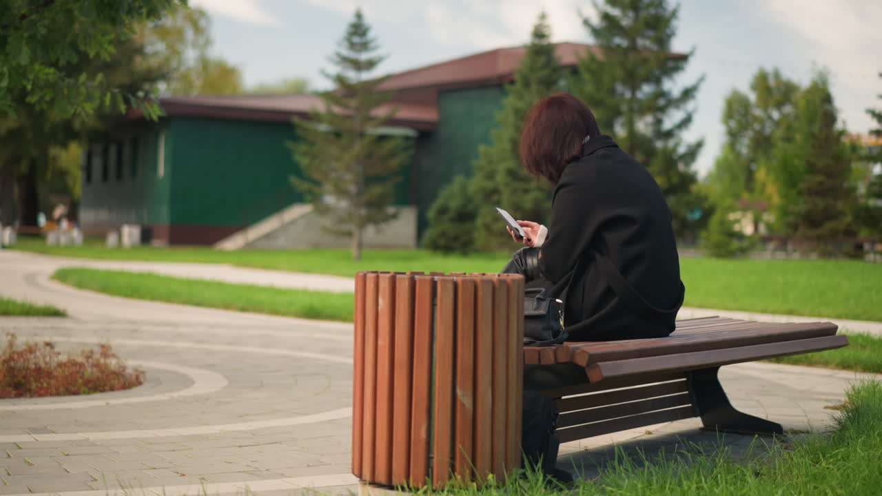 vista trasera de una mujer vestida con abrigo y botas negras, sentada en un banco del parque usando un teléfono inteligente, rodeada de exuberante vegetación, caminos curvos y un ambiente al aire libre sereno en un parque urbano