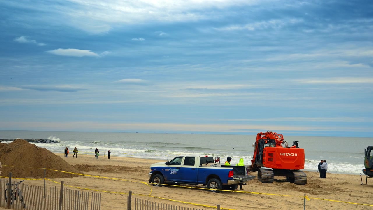 Nueva York, EE. UU., 1 de abril de 2026: Vista trasera de excavadora naranja y camión en la playa. Parte trasera de una excavadora pesada y una camioneta pickup azul en una playa de arena con el océano de fondo.