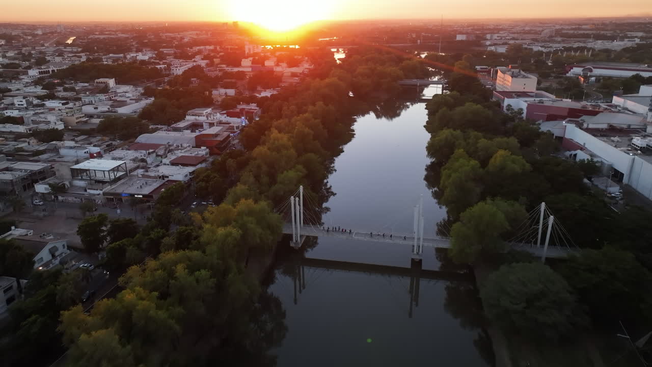 vista aérea hora dorada sobre un río con puente peatonal y panorama de metrópolis en el horizonte