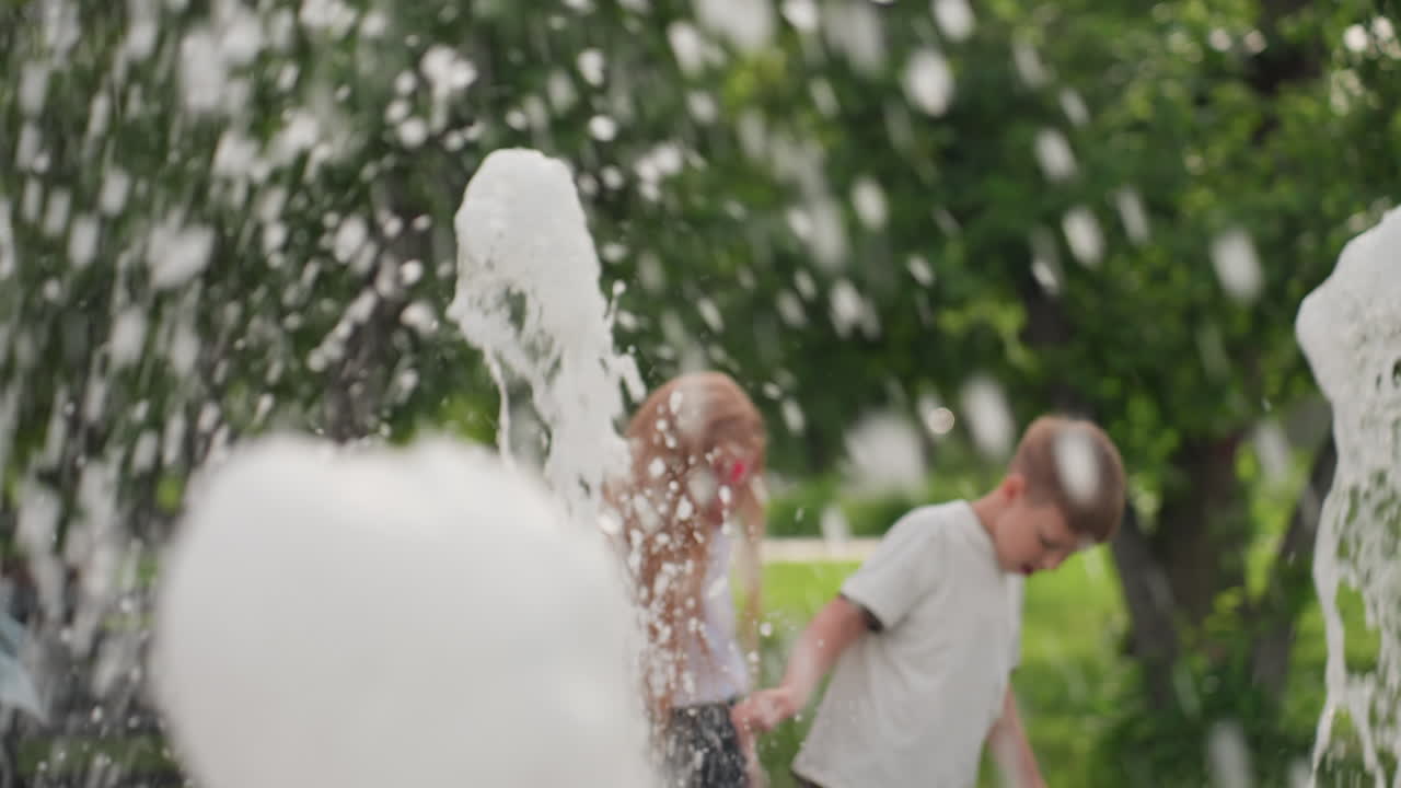 Energetic children chatting and playing around recreational park fountain, water jets splashing, blurred greenery in background, summer day showing movement and joyful interaction