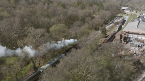 Aerial-drone-shot-of-a-steam-train-engine-moving-past-a-factory