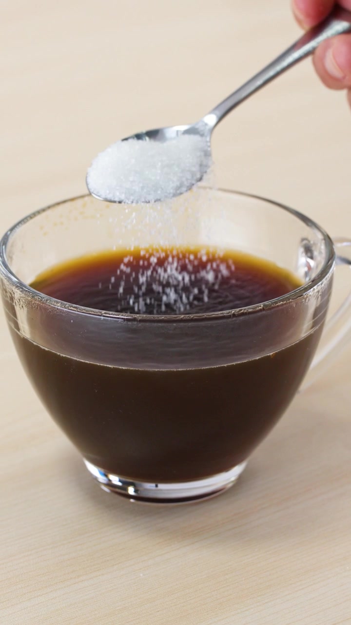 Zucchero granulato cade da un cucchiaio di metallo in una tazza di vetro di caffè nero.
