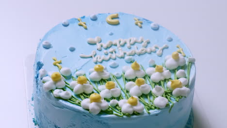 Zooming-out-of-a-close-up-shot-of-a-blue-frosted-birthday-cake,-decorated-with-small-white-flowers-with-yellow-tips-and-green-accents