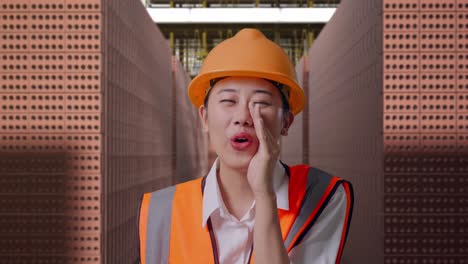 close up of asian female engineer with safety helmet yelling with hand over mouth while standing with red brick packed in stacks are stored