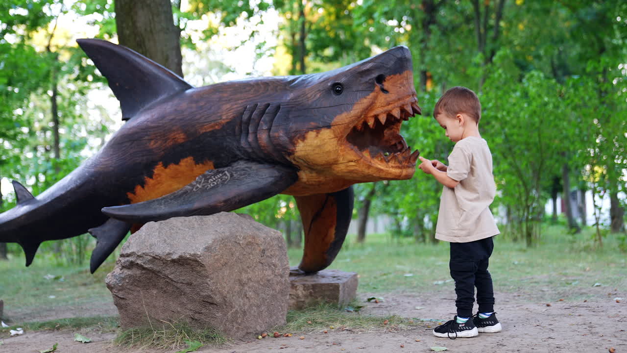 Cute baby boy comes up to a wooden sculpture of shark. Kid puts acorn into the sculpture jaws.