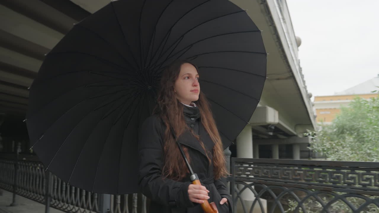 Donna caucasica che cammina sotto un ombrello nero, lunghi capelli ricci, cappotto scuro, ombrello vintage con goccioline, in piedi sotto un sottopassaggio di cemento, ringhiera in ferro battuto, luce fioca e malinconica, atteggiamento contemplativo