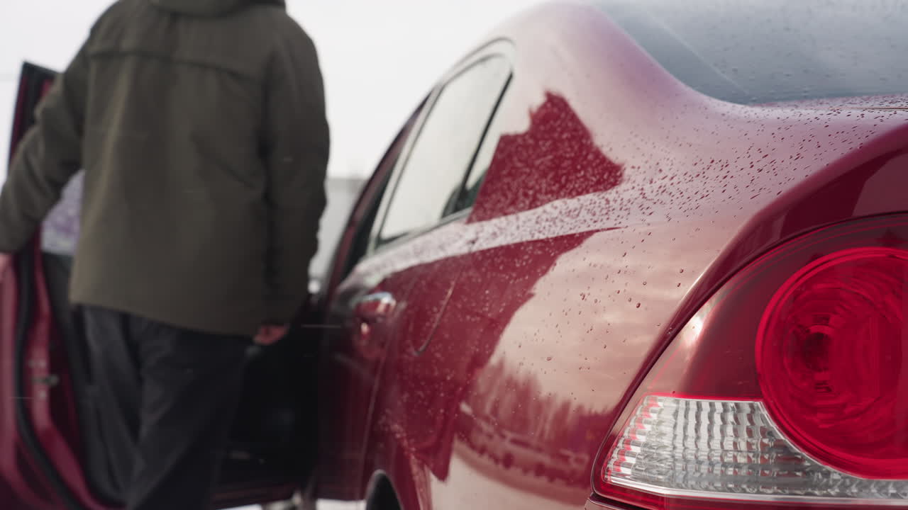 Rear side view of red car covered with water droplets as businessman in winter jacket opens door and enters to drive, with surrounding parked cars reflected on glossy surface in snowy outdoor setting